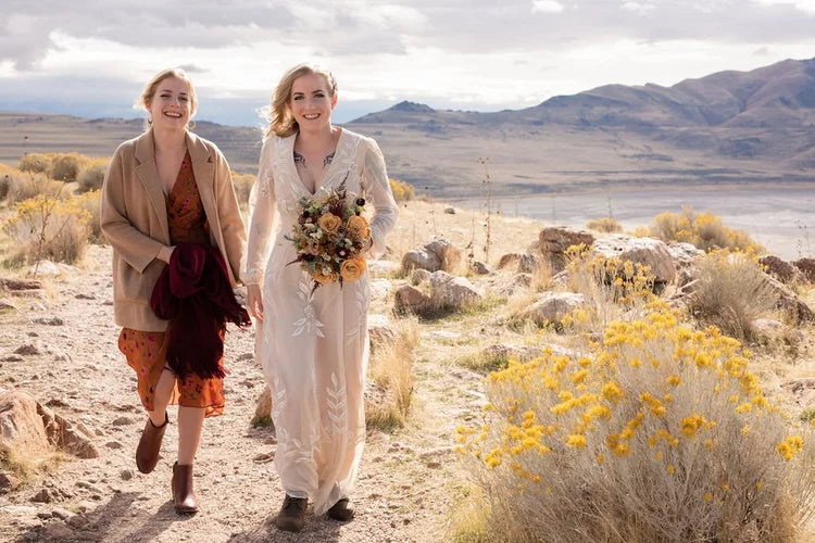 Two women walking in a desert landscape with mountains in the background carrying Apiana Blooms wedding bouquet